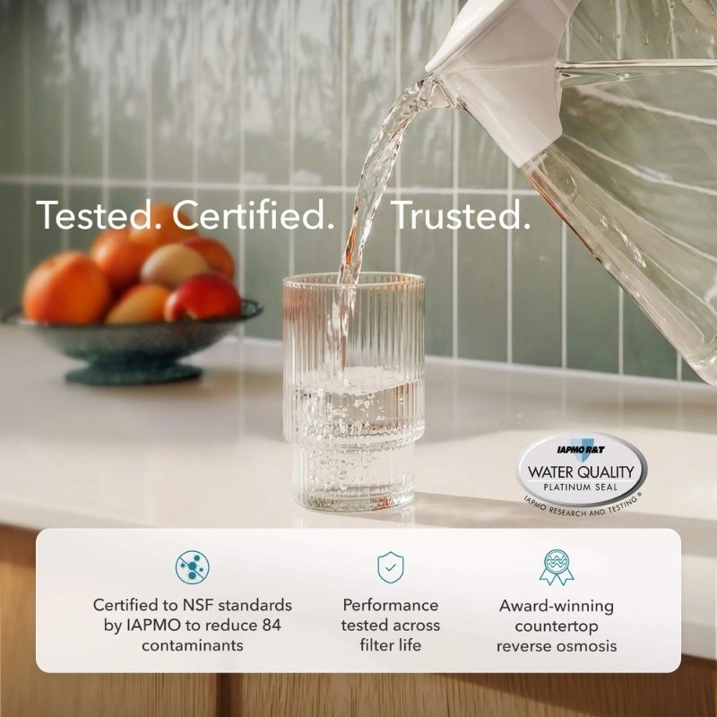Water being poured from AquaTru Carafe - Glass into a glass on a kitchen counter with IAPMO water quality certification seal.