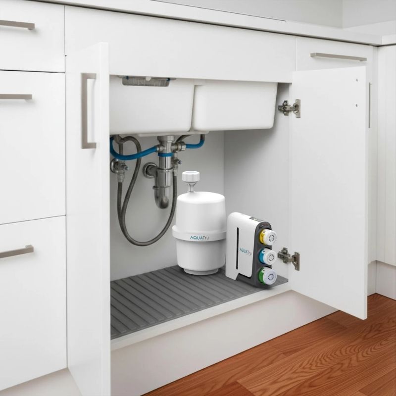 A kitchen sink under-cabinet water filtration setup shown installed inside an open white cabinet. Visible are a white AquaTru Under Sink Reverse Osmosis Water Purifier - Gen2, a white and gray faucet filter cartridge, and plumbing pipes behind them on a gray rubber tray.