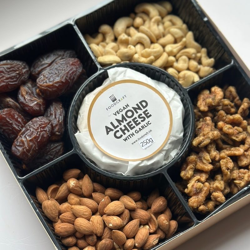 A gourmet snack box (Artisanal Gluten Free Vegan Cheese & Sprouted Nuts Platter - Almond Garlic Cheese
Foodcraft Online Store) featuring a 250g round of vegan almond cheese with garlic in the center, surrounded by five compartments of nuts and dates: cashews, almonds, walnuts, and dates.