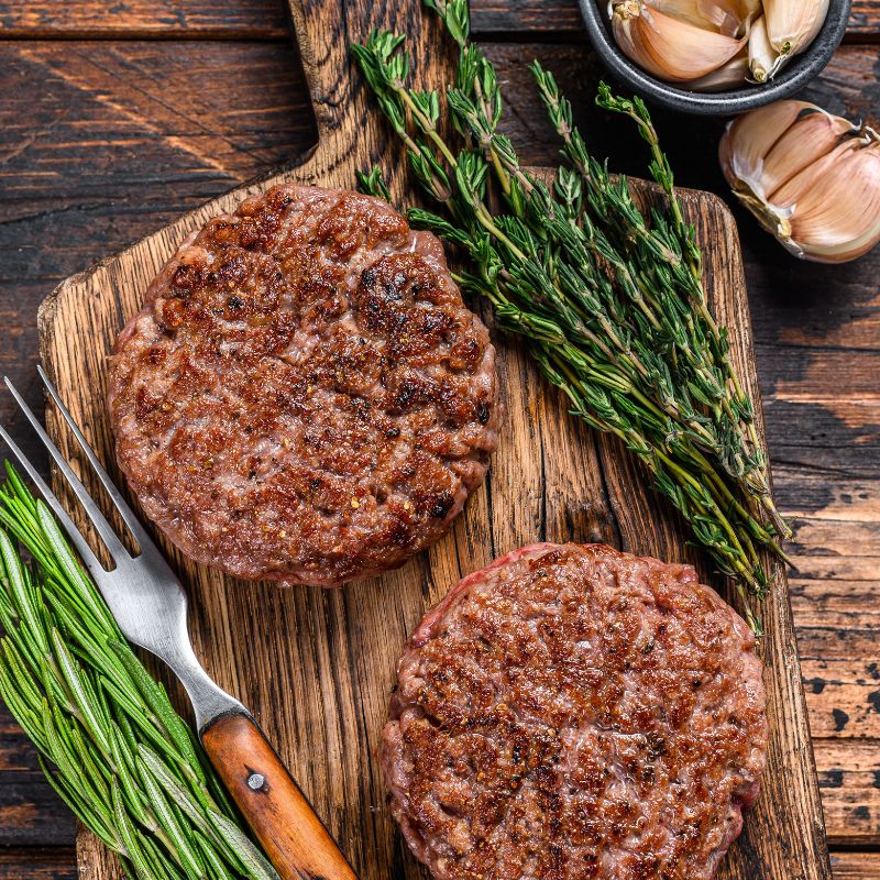 Two Australian AMG Grass Fed Burger Patty on a wooden cutting board with herbs and garlic cloves.