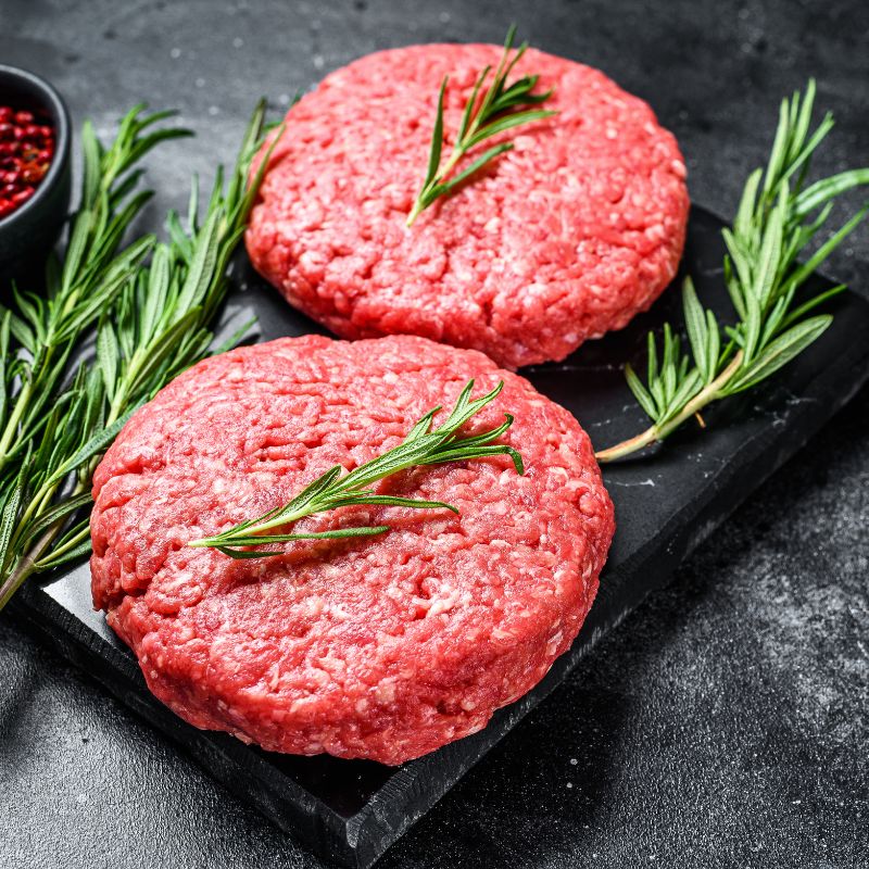 Two raw Australian AMG Grass Fed Burger Patties on a black slate board with rosemary sprigs.