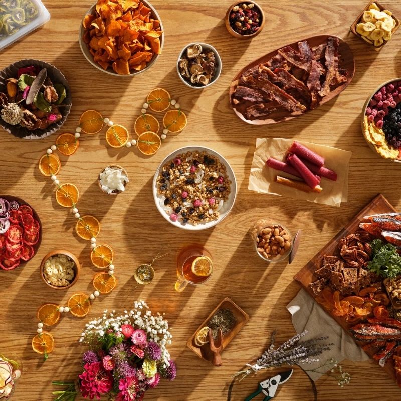 An overhead styled flat-lay of assorted gourmet snacks and foods  made with Excalibur 10 Tray Performance Digital Dehydrator, in Stainless Steel - DH10SSSS13 arranged on a wooden table, including dried fruit, nuts, crackers, a bowl of granola, flowers, and a drink, with no people or real environment beyond the tabletop setup.