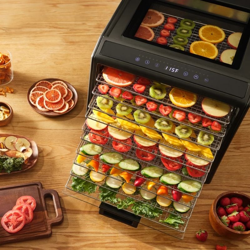 Excalibur 10 Tray Performance Digital Dehydrator, in Stainless Steel - DH10SSSS13 with multiple clear trays loaded with sliced strawberries, kiwi, oranges, apples, cucumbers, and greens, shown on a wooden table with bowls of fresh produce around it.