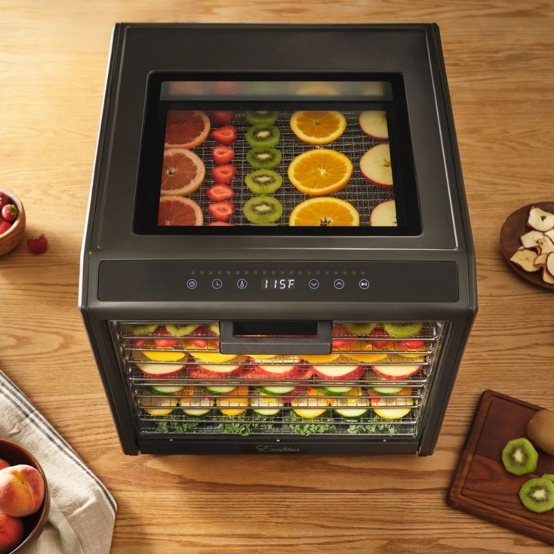 A countertop Excalibur 10 Tray Performance Digital Dehydrator, in Stainless Steel - DH10SSSS13 with a black exterior and glass door, shown on a wooden table with sliced fruits inside. The brand name 'Midea' is visible on the lower front.