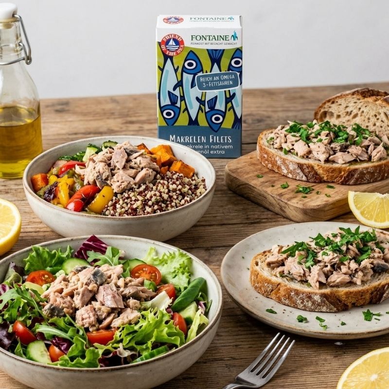 Fontaine Chub Mackerel Fillets in Organic Olive Oil is shown in a kitchen setting with prepared meals. A box of Fontaine Chub Mackerel Fillets in Organic Olive Oil - 90g is standing behind bowls of salad and toasted bread on a rustic wooden table, with a bottle of olive oil and lemon slices nearby.