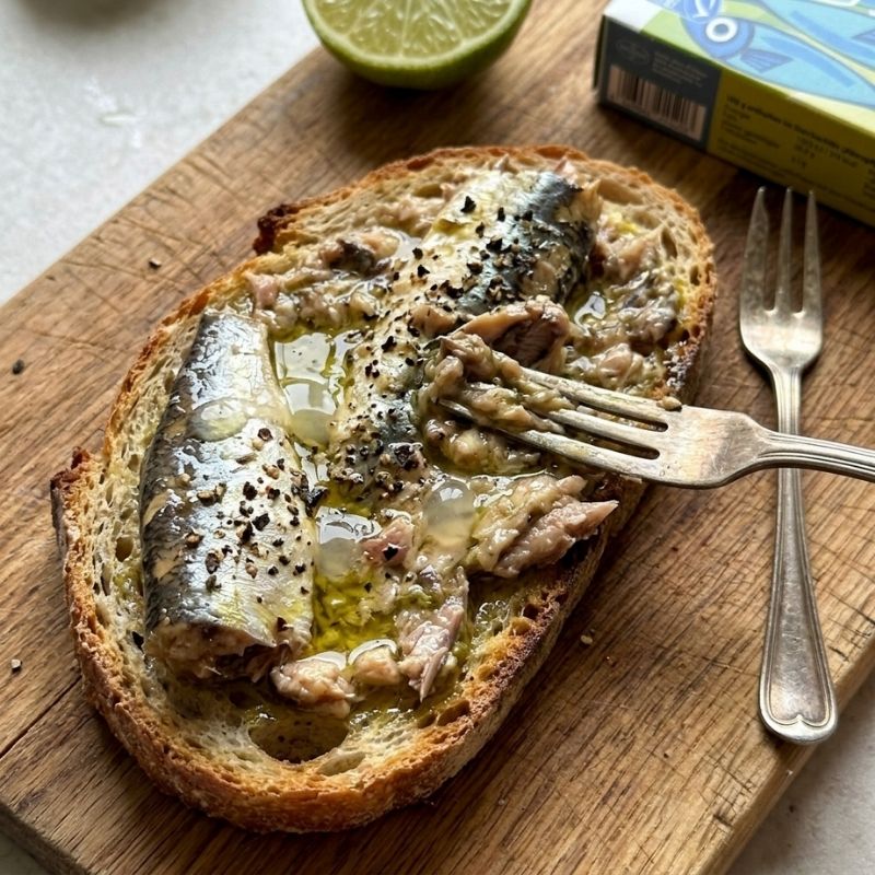 A close-up of a piece of toast topped with sardines, oil, and black pepper, resting on a wooden cutting board with a fork, a lime half, and a box of Fontaine Sardines in Organic Olive Oil - 120 g in the background.