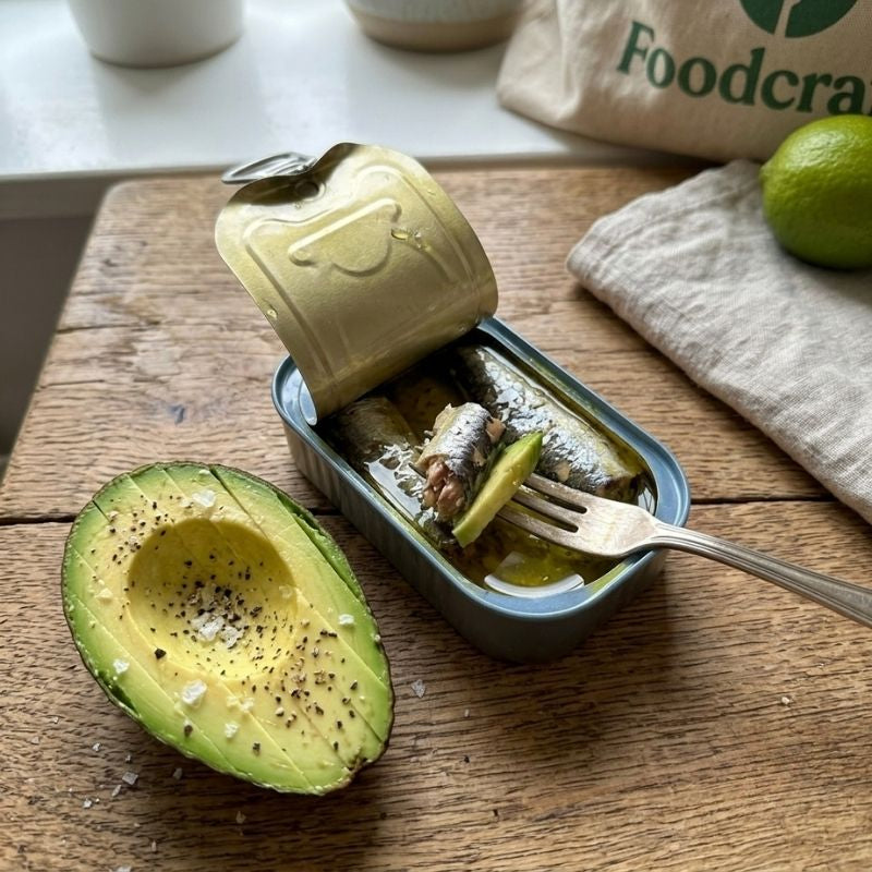 An open can of Fontaine Sardines in Organic Olive Oil - 120 g on a rustic wooden cutting board, with a fork lifting a piece of fish. A halved avocado seasoned with sea salt sits beside it, and a beige tote bag with the green 'Foodcraft' logo is visible in the background.