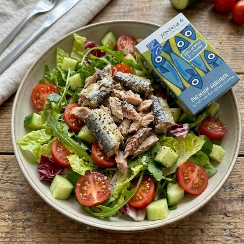 A plated salad on a wooden table with sliced sardines on top, tomatoes, cucumbers, greens, and a box of Fontaine Sardines in Organic Olive Oil - 120 g. The box is blue and yellow with fish illustrations.