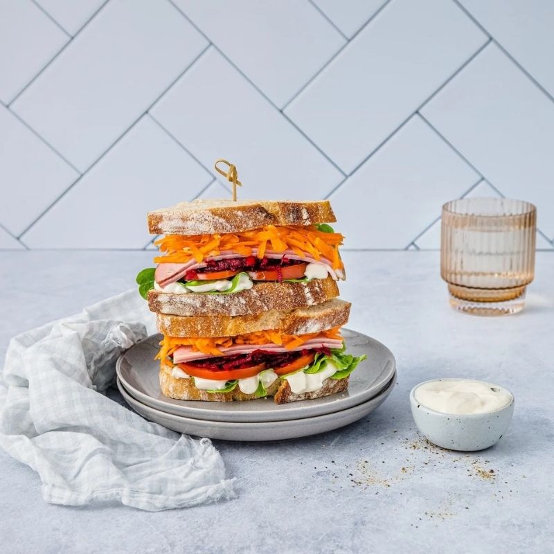 A large stacked sandwich on two gray plates, filled with shredded carrots, tomatoes, greens, and meats, set on a light gray surface with a small dish of Gevity Body Glue Original Great Guts Mayo and a glass in the background.