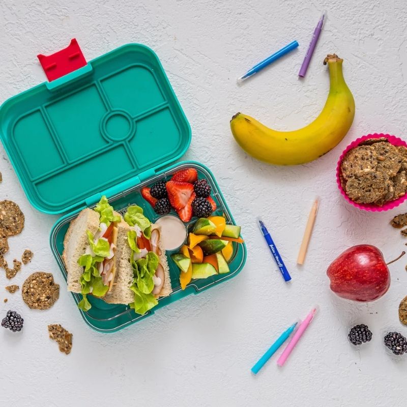 A teal silicone bento lunch box with a red latch, shown open and filled with a sandwich with Gevity Body Glue Original Great Guts Mayo, fruit, and a small dessert. The lunch box is photographed in a styled flat-lay arrangement on a white textured background with a banana, apple, berries, cookies, and colored pencils around it.