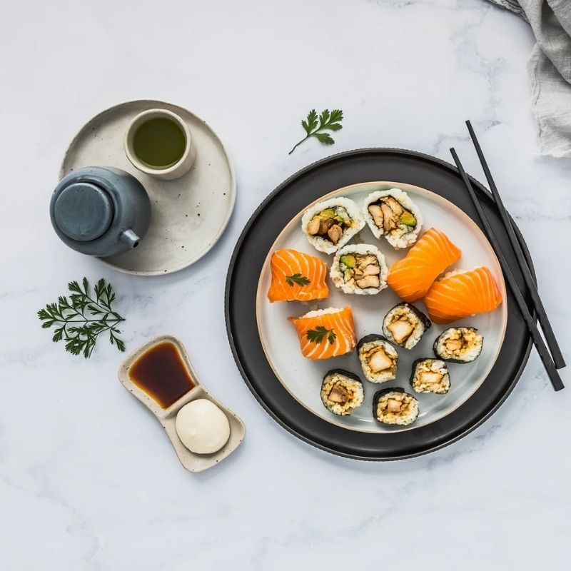 Assorted sushi on a plate with a cup of green tea, soy sauce, and Gevity Body Glue Original Great Guts Mayo on a light gray surface.
