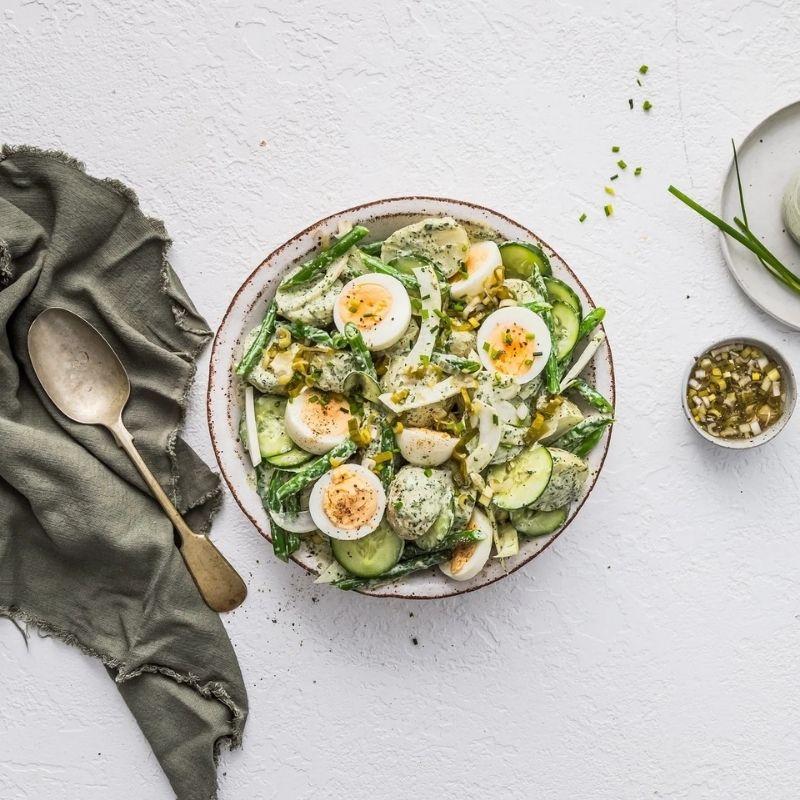 A top-down view of a bowl of salad on a textured white surface, featuring mixed greens, cucumber slices, hard-boiled egg halves, and a creamy Gevity Body Glue Original Great Guts Mayo dressing. A dark green cloth napkin with a spoon is on the left, and a small bowl of garnish is on the right.