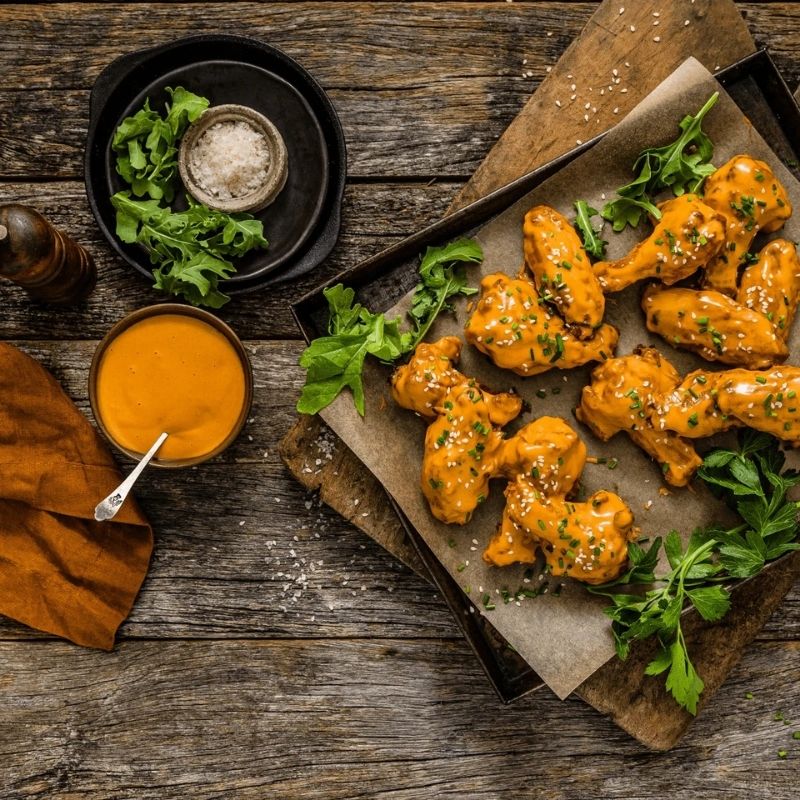 Orange buffalo-style chicken wings on a wooden serving board with parchment paper, garnished with chopped chives and sesame seeds, set on a rustic wooden table with Gevity Body Glue Smoky BBQ Great Guts Mayo, greens, and a pepper mill.