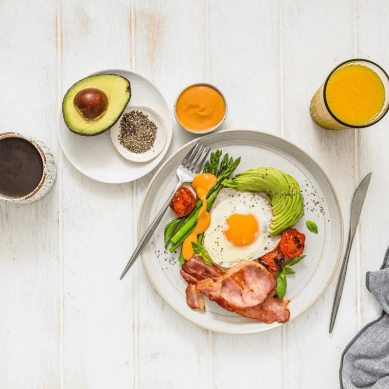 A top-down view of a breakfast spread on a white wooden table, featuring a plate with a fried egg, bacon, avocado, asparagus, tomatoes, and Gevity Body Glue Smoky BBQ Great Guts Mayo, alongside an avocado half, a small bowl of seeds, a glass of orange juice, and a cup of coffee.