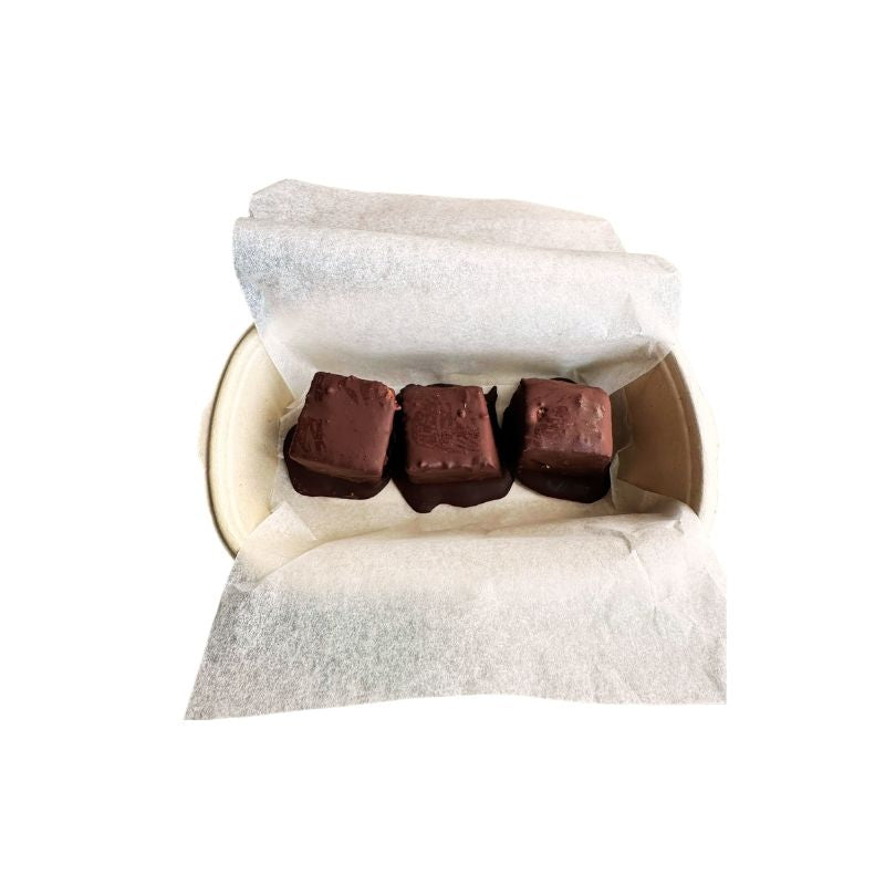 Three chocolate-covered Gluten Free Keto Dark Chocolate Coated Lemon Poppy Seed Cake - 150g pieces arranged in a paper box lined with white parchment paper, photographed on a plain white background.