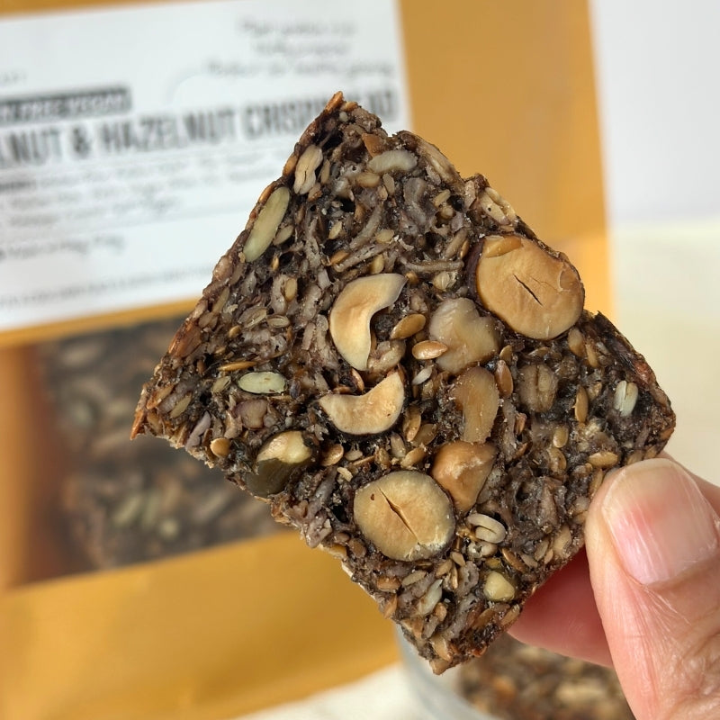 A hand holding a square piece of  Gluten Free Walnut & Hazelnut Crispbread - 180g
Foodcraft Online Store with a blurred package in the background.