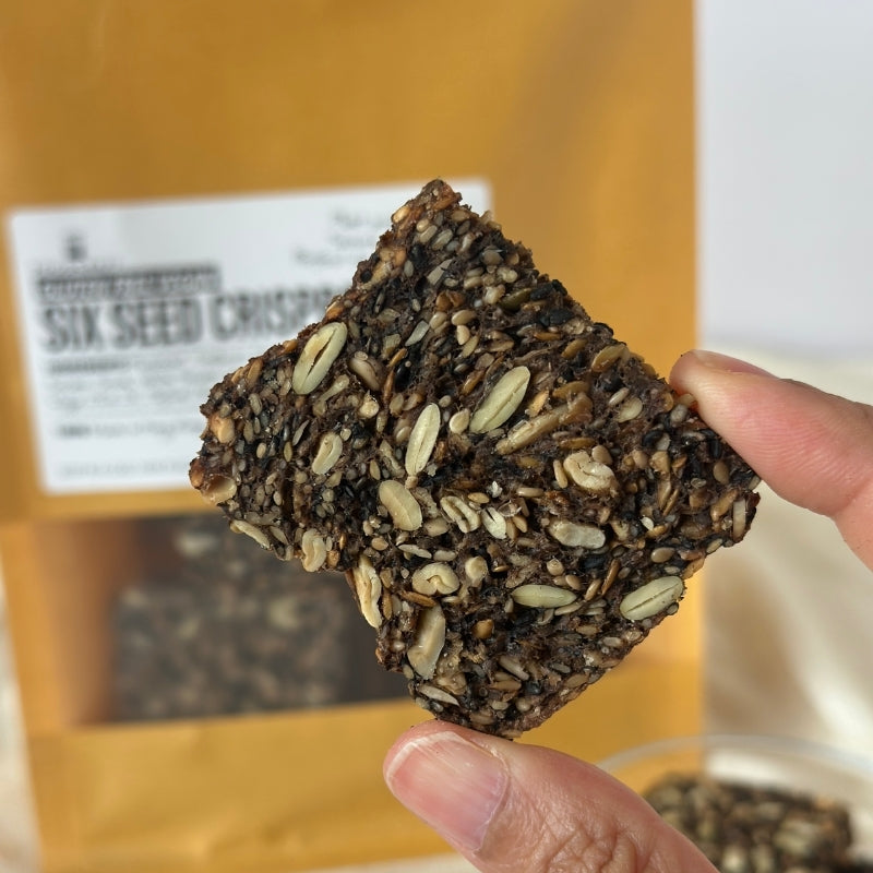 A hand holding a square piece of sGluten Free 6 Seed Crispbread - 180g in front of a brown bag of Gluten Free 6 Seed Crispbread - 180g