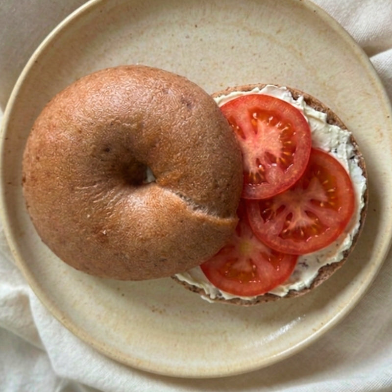 A close-up of a Gluten-Free Soft Original Sourdough Bagels (from Foodcraft Online Store)  half filled with cream cheese and sliced red tomatoes on a light beige plate.