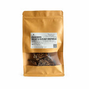 A stand-up kraft paper pouch of Gluten Free Walnut & Hazelnut Crispbread - 180g, featuring a clear window at the bottom that displays the Gluten Free Walnut & Hazelnut Crispbread - 180g pieces inside. The packaging is a warm golden-brown color with a white label in the center.