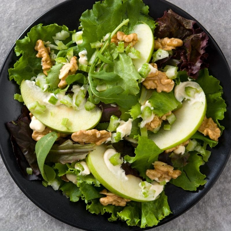 A top-down view of a WALDORF salad made with Granny Smith Apples (Green Apples)  on a black plate, featuring mixed green and purple lettuce, creamy dressing, and chopped celery.