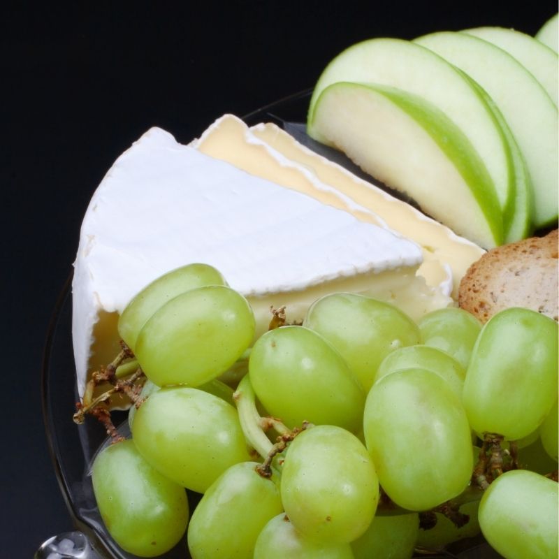 A close-up of a cheese board with wedges of brie and camembert, sliced green apples, green grapes, and a piece of bread on a dark plate against a black background.