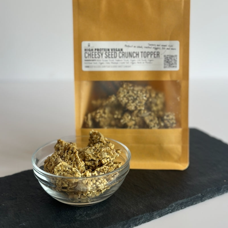 A kraft paper pouch labeled 'High Protein Vegan Cheesy Seed Crunch Topper' stands behind a small clear glass bowl filled with the A stand-up kraft paper pouch  High Protein Vegan Cheesy Seed Crunch Topper - 100g (Foodcraft Online Store) The pouch is centered on a clean white background with a soft shadow on a dark slate surface against a light neutral background.