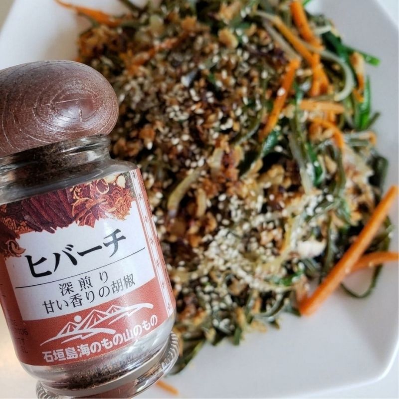 A glass bottle of Japanese MSG-Free Hibachi Pepper Seasoning with a dark wooden cap and a brown-and-white label, shown in the foreground next to a prepared salad on a white plate.