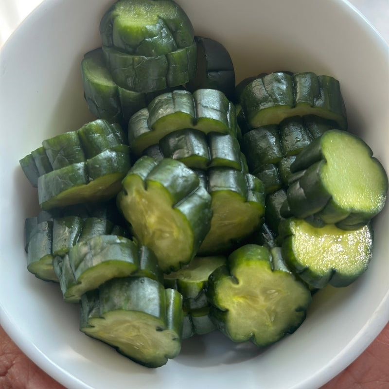 Sliced Living Probiotic Nukazuke Cucumber Pickles - 160g with dark rinds piled in a white bowl, with a hand holding the bowl at the bottom edge.