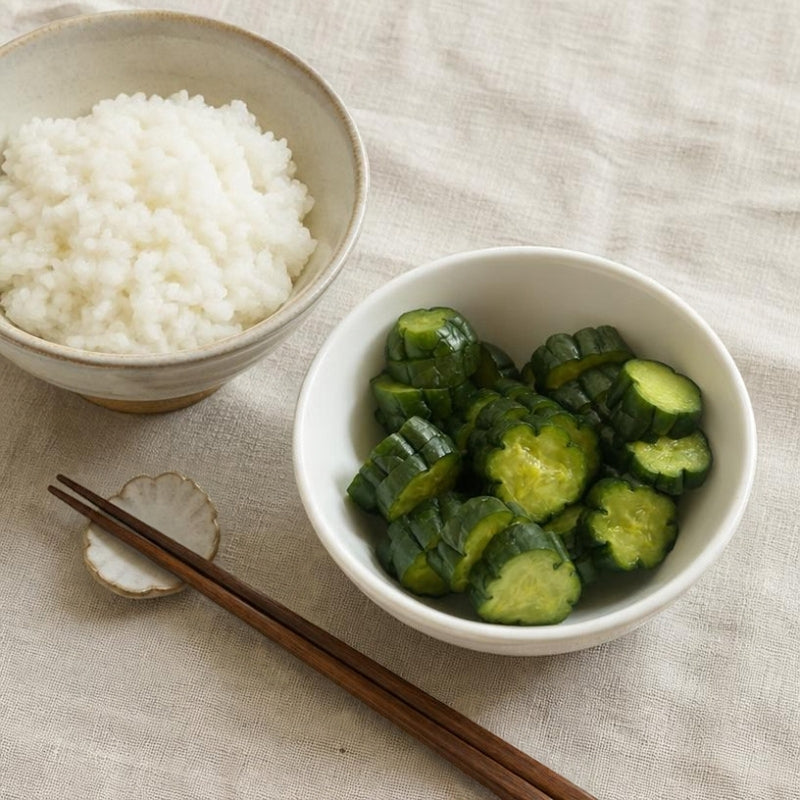 A styled food scene on a beige linen tablecloth showing a white bowl of sliced Living Probiotic Nukazuke Cucumber Pickles - 160g and a beige bowl of white rice, with brown chopsticks resting on a small dish.