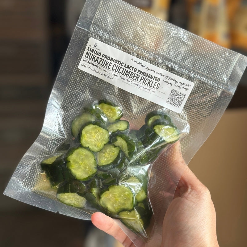 A hand holding a clear vacuum-sealed bag of sliced cucumbers with a white label reading 'Living Probiotic Lacto Fermented Nuzake Cucumber Pickles.'.