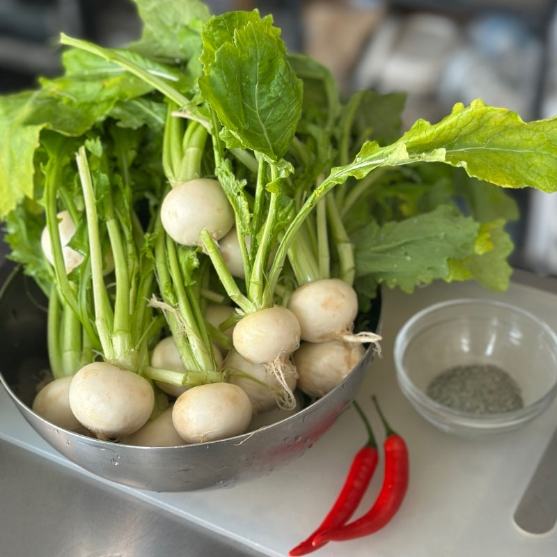 Kabu Turnips with green leaves in a metal bowl on a kitchen counter with red chilies and a small container of salt.