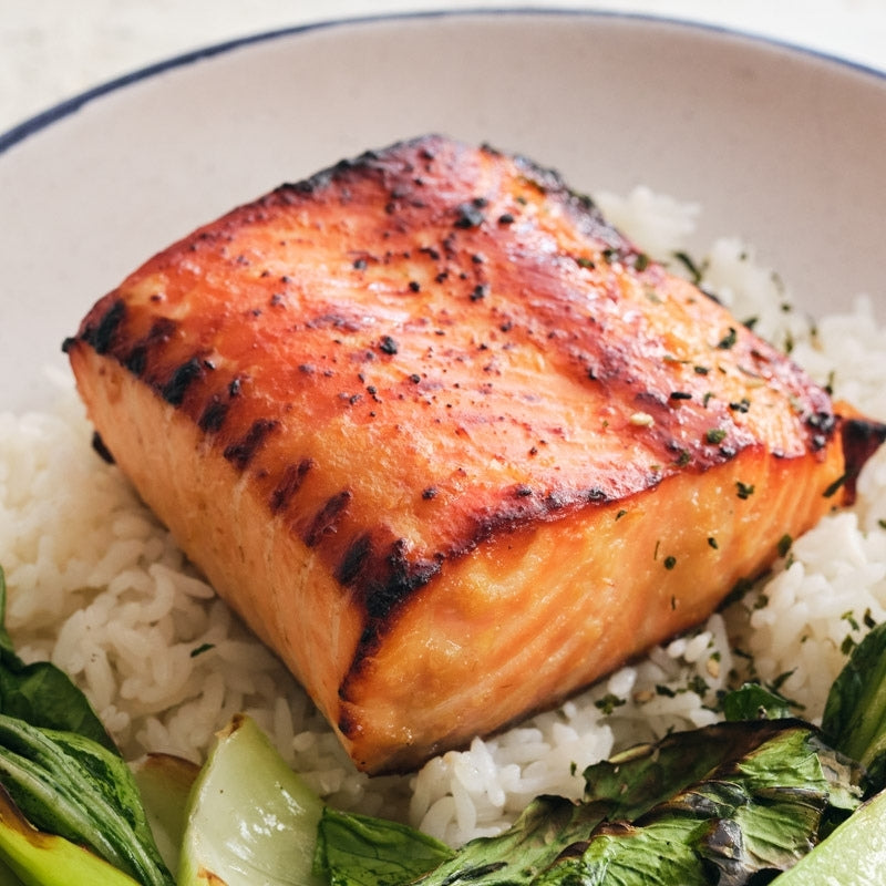 A close-up of a grilled salmon fillet made with Living Probiotic Chili Miso Dressing - 220g
Foodcraft Online Store  resting on a bed of white rice with green vegetables in a light-colored bowl