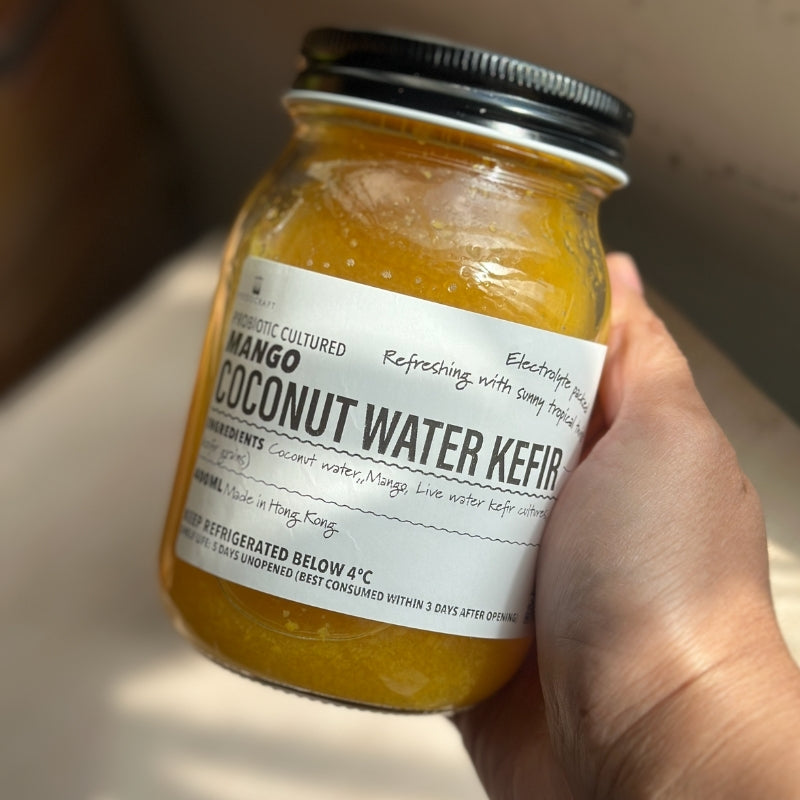 A hand holding a glass jar of Mango Coconut Water Kefir - 400ml
Foodcraft Online Store. The jar has a black metal lid and a white label with black text, set against a warm, softly blurred background.