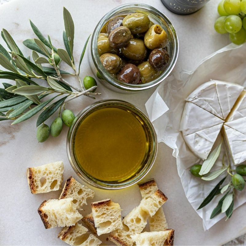 Mazza Olio Extra Virgin Olive Oil, olives, bread, and cheese on a marble surface with olive branch garnish.