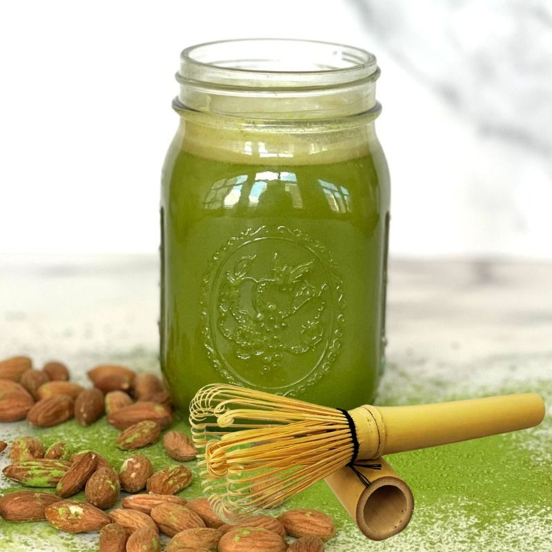 Glass jar with almond matcha latte, almonds, and a bamboo whisk on a white background