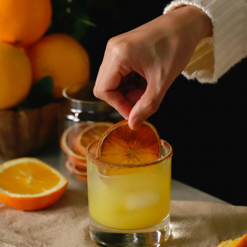 Hand placing a dried organic orange slice into a glass of yellow liquid with oranges in the background.