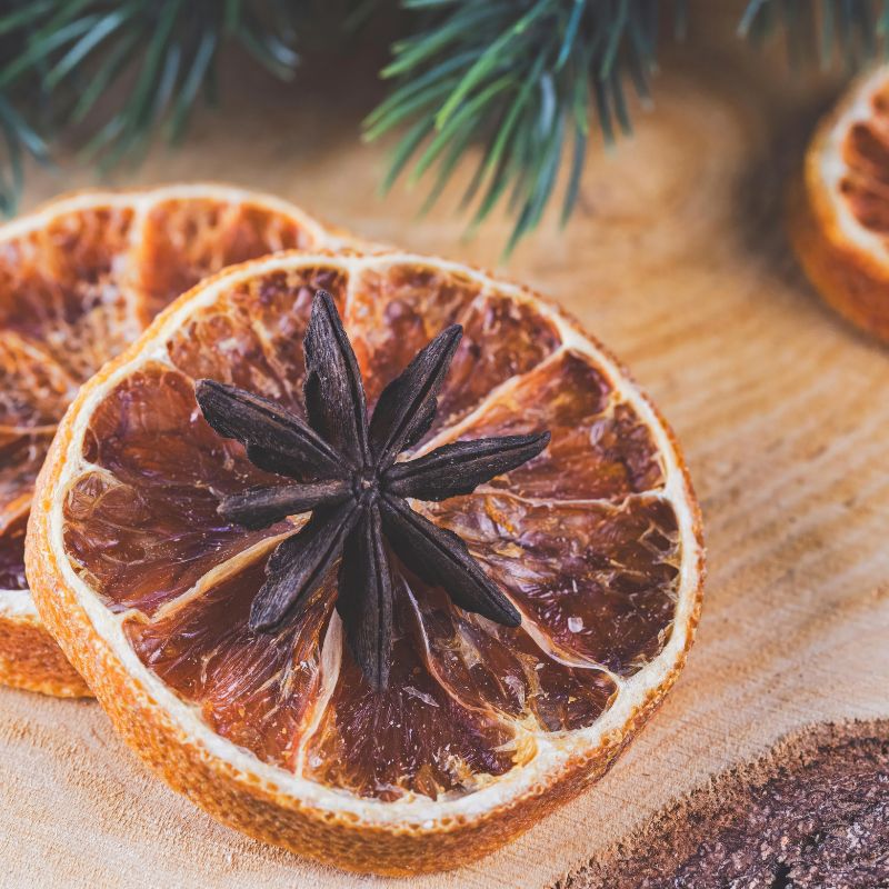 Dried organic orange slices with a star anise on a wooden surface, surrounded by pine branches.