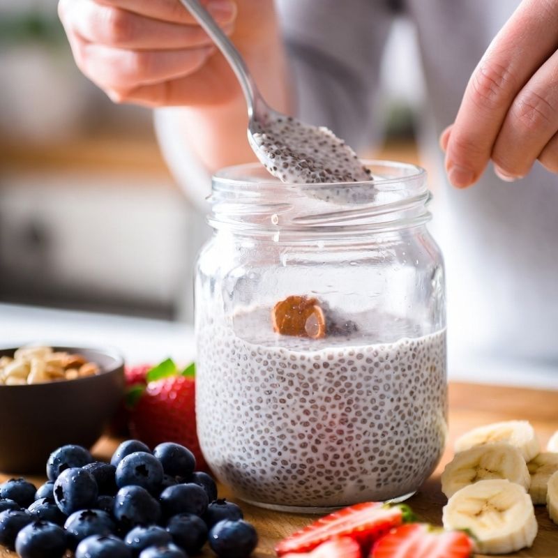 Glass jar of chia pudding with a spoon, surrounded by fruits and granola on a wooden surface.