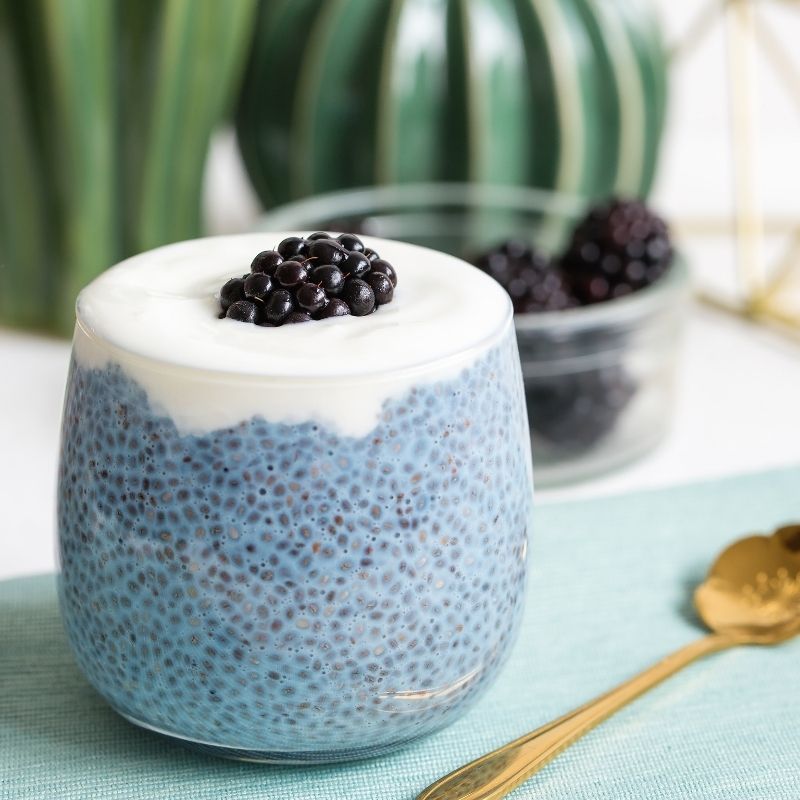 Glass container of blue chia pudding with white cream and black berries on a light green surface.