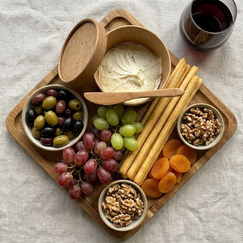 A wooden charcuterie board arranged on a linen surface with a bowl of Organic Vegan Spreadable Garlic & Herbs Cream Cheese, a spoon, crackers, olives, grapes, apricots, walnuts, and a glass of red wine.