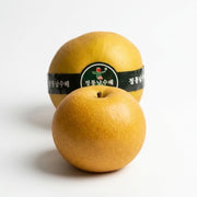 Two golden-yellow Nashi Pears from Korea on a clean white background, with one pear in the foreground and another behind it wrapped in a black label with Korean text and a small green logo.