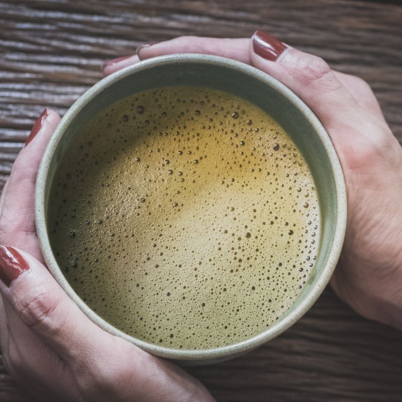 Hands holding a bowl of frothy Pure Hojicha (Unsweetened)  on a wooden surface