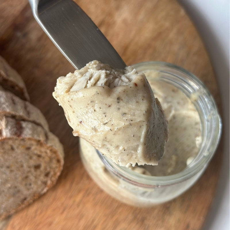 A close-up of a knife lifting a scoop of Raw Vegan Butter with Herbs- 180g
(Foodcraft Online Store) from a small clear glass jar, with sliced bread on a wooden board in the background.