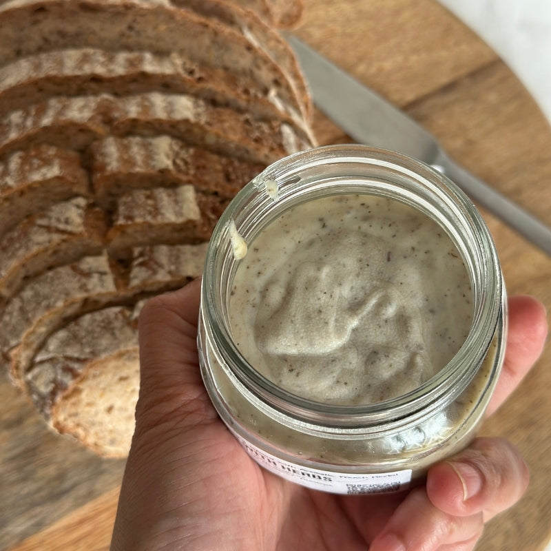 A hand holding an open glass jar of Raw Vegan Butter with Herbs- 180g
(Foodcraft Online Store) with sliced rustic bread and a knife blurred in the background on a wooden surface.