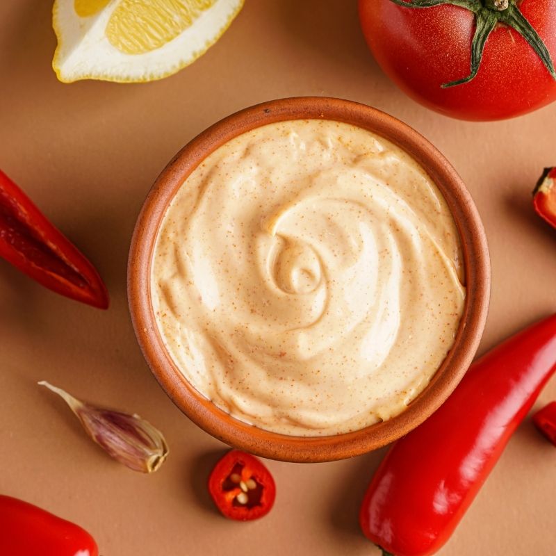 Bowl of creamy sauce surrounded by Red Jalapeno Peppers, a lemon, and a tomato on a brown background