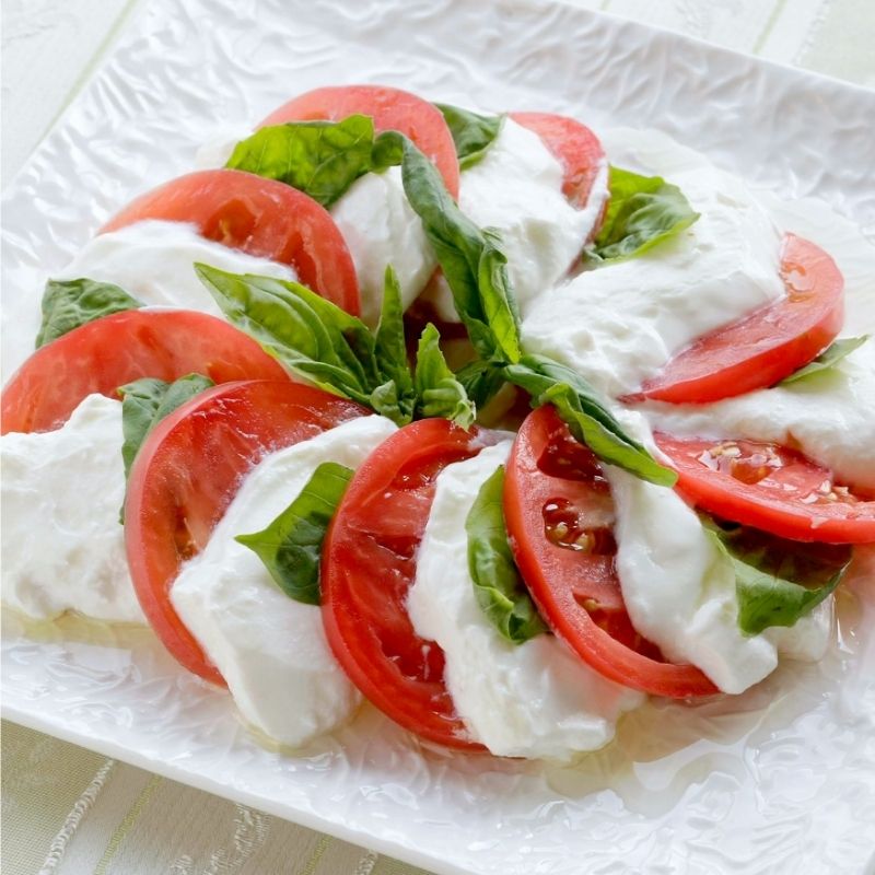 Caprese salad with sliced red tomatoes, fresh green basil leaves, and creamy white mozzarella cheese, arranged on a white square plate. the cheese was made in Stainless Steel Greek Yogurt Strainer 希臘優格水濾器.
