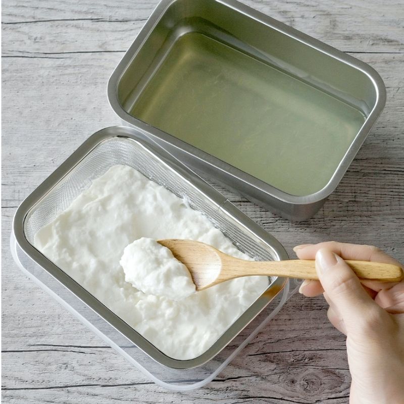 Stainless Steel Greek Yogurt Strainer 希臘優格水濾器 with a clear lid and mesh filter, shown with white yogurt or cream inside and a wooden spoon being used to scoop. Another empty container sits behind it on a gray wood-textured surface.