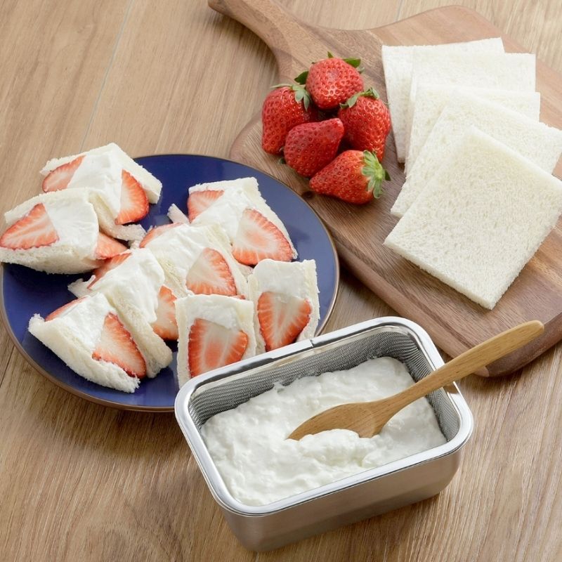 Stainless Steel Greek Yogurt Strainer 希臘優格水濾器 filled with white yogurt, with a wooden spoon inside, set on a wooden table. A blue plate with strawberry sandwich slices and sliced white bread on a wooden cutting board are arranged nearby.