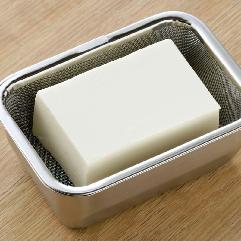 A silken tofu resting inside Stainless Steel Greek Yogurt Strainer 希臘優格水濾器 with a mesh liner, set on a light wood grain surface.