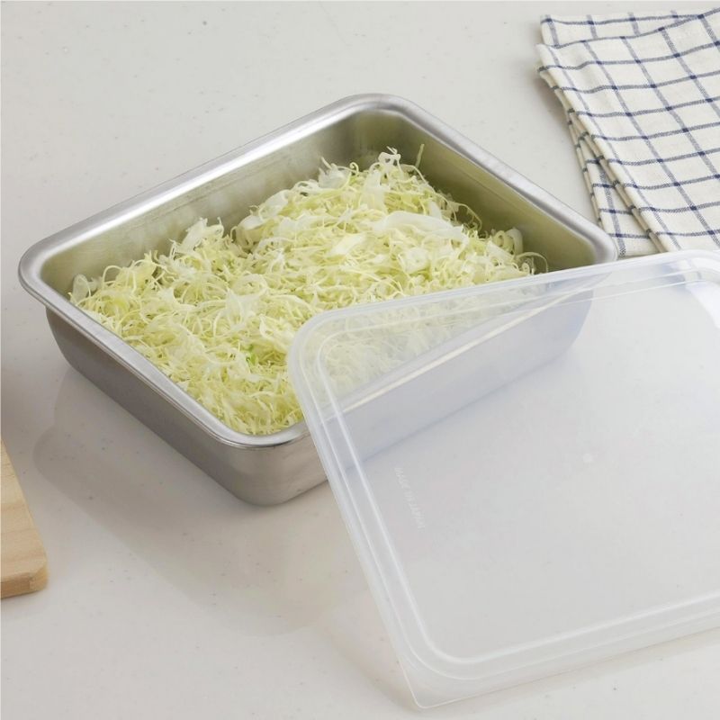 Stainless Steel Storage Prep Tray with Lid (Wide Deep Size) filled with shredded pale green cabbage, shown with a clear plastic lid resting in front on a light countertop. A blue-and-white checkered cloth is visible in the background.